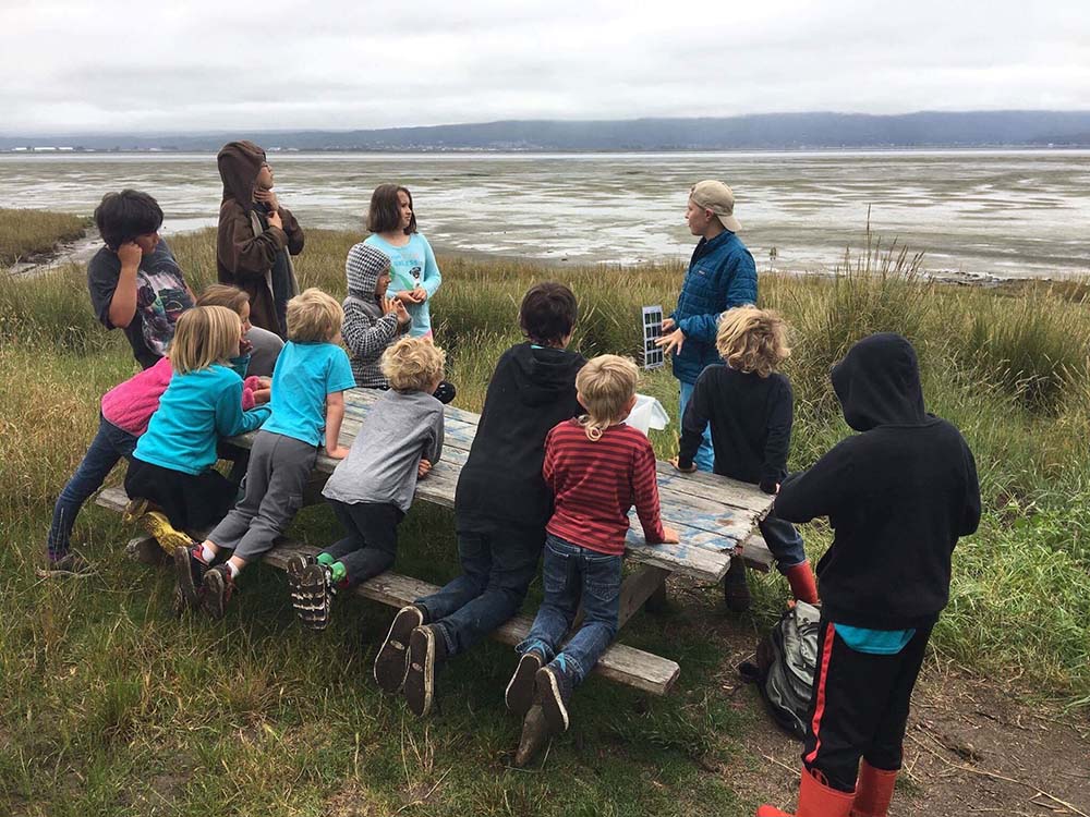 A group of kids are at a picnic table on a hill overlooking the bay. They are watching an instructor, who is speaking and holding a photo.